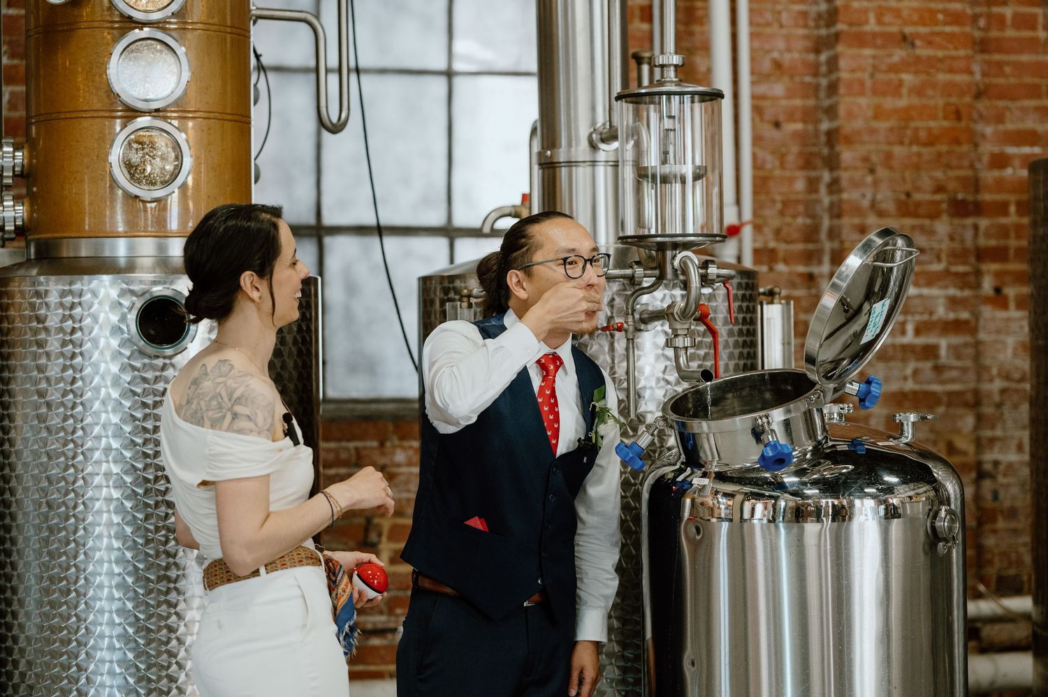 Wedding couple examining stainless steel brewing equipment in an industrial distillery setting.