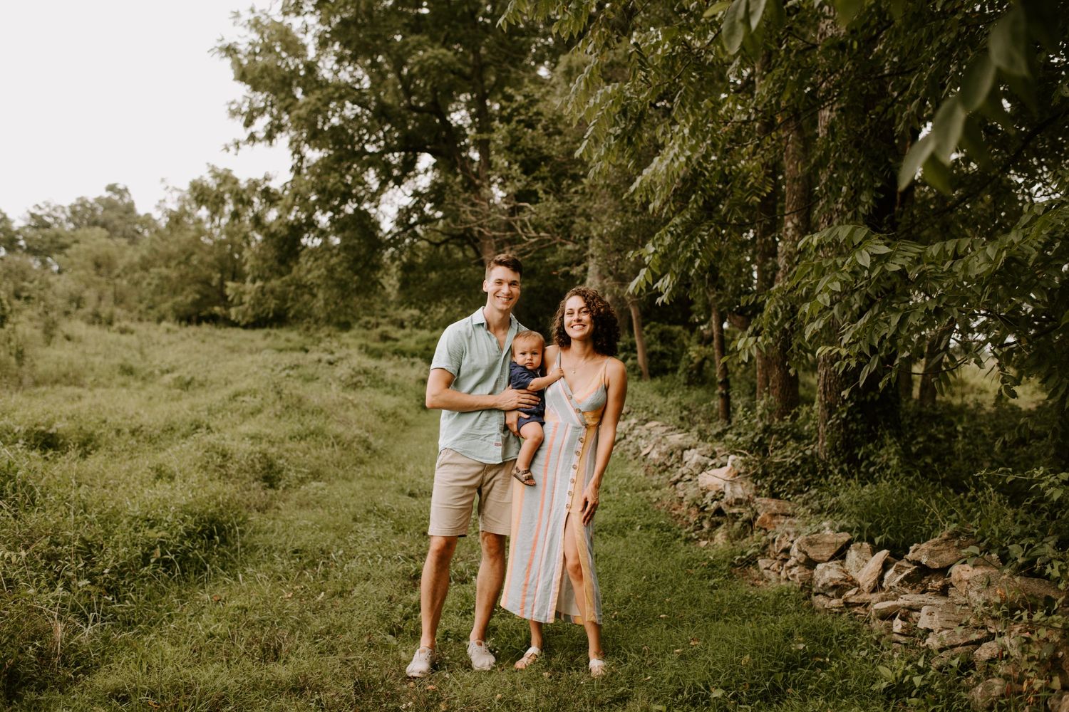 Family standing together on grassy hillside near stone wall during outdoor photo session.