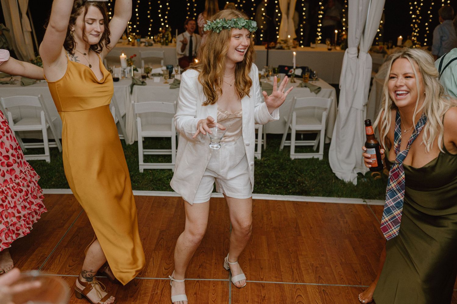Wedding party members dance on a wooden floor under string lights.