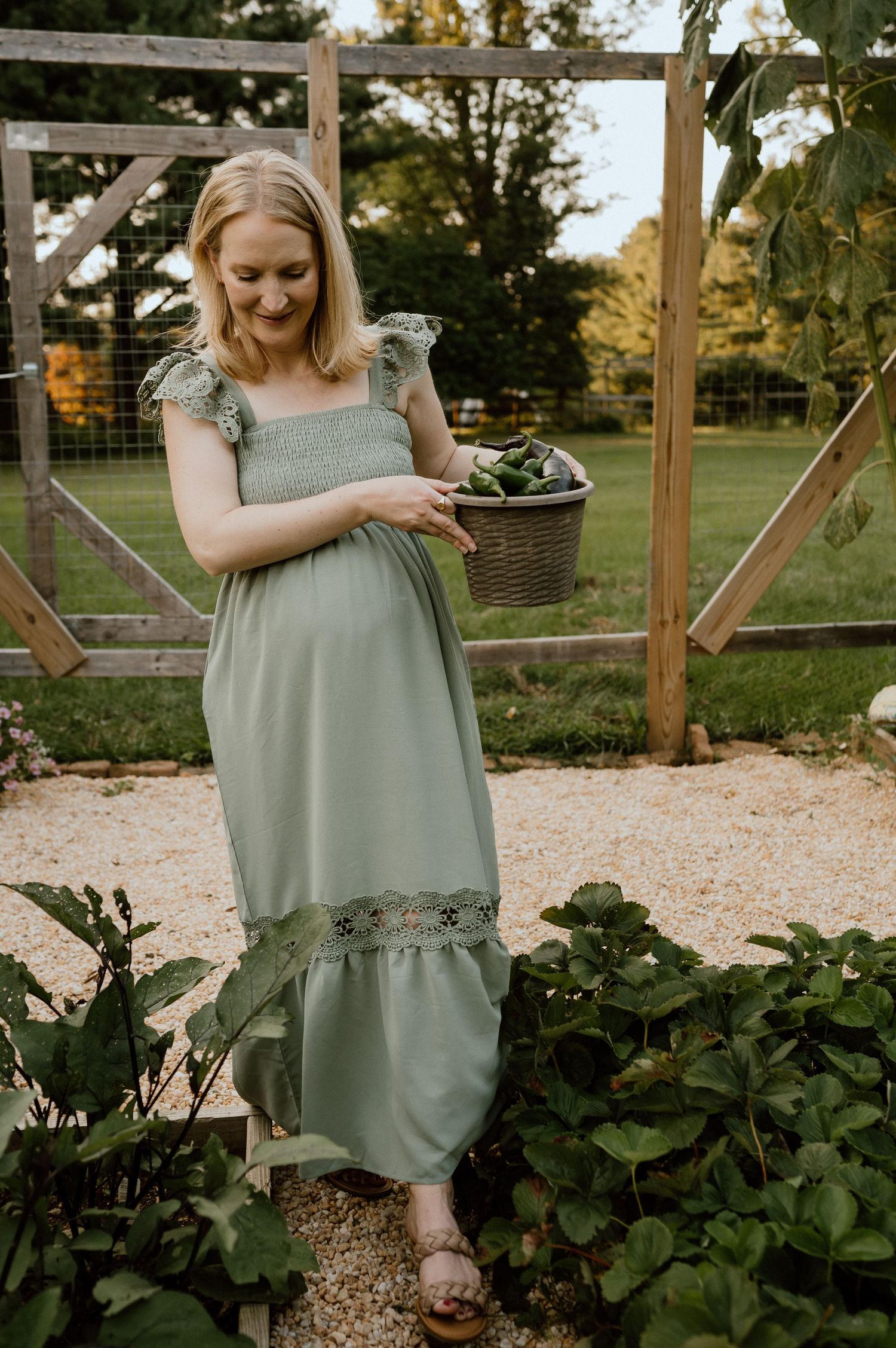 Person in sage green maxi dress standing by garden gate holding basket of vegetables.