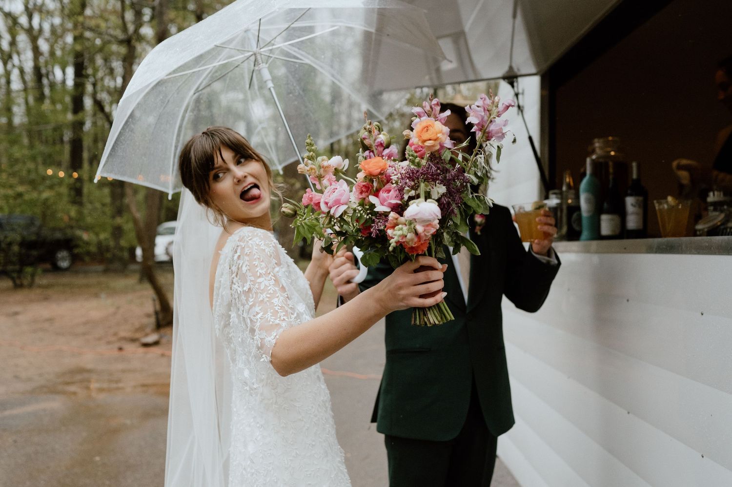 A joyful couple laughs while holding a colorful bouquet under a clear umbrella at their outdoor wedding.