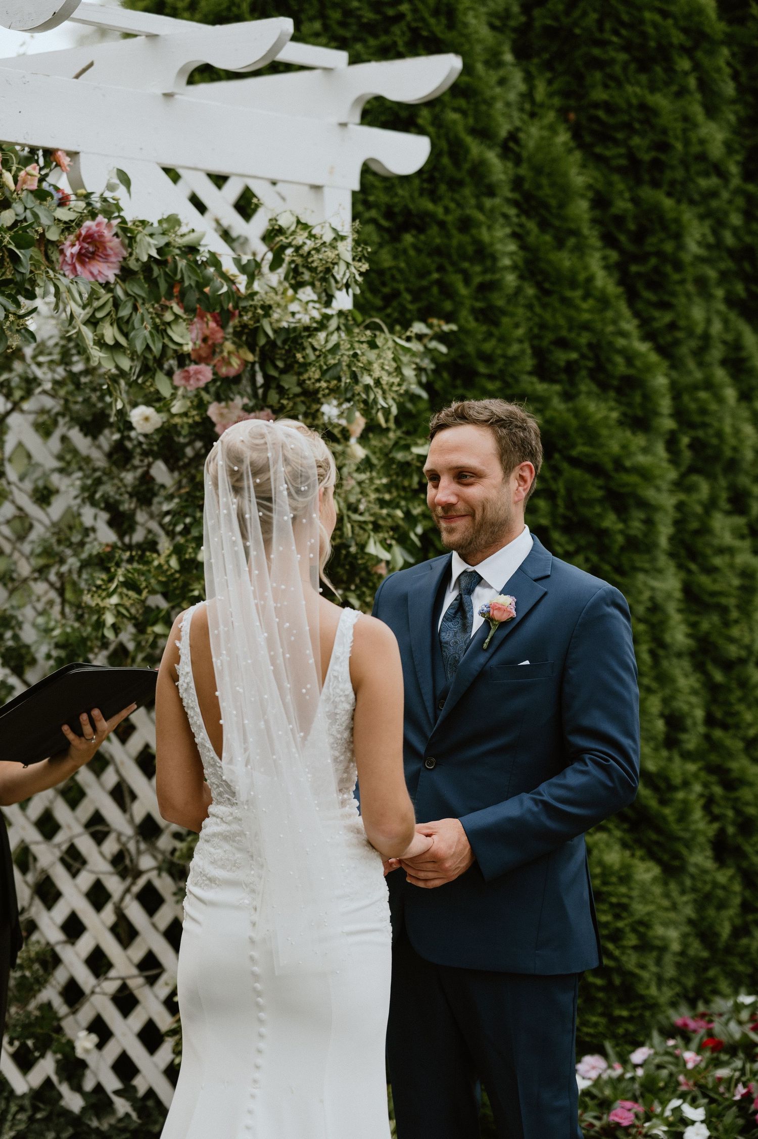A wedding ceremony takes place under a white lattice arch decorated with pink roses and greenery.