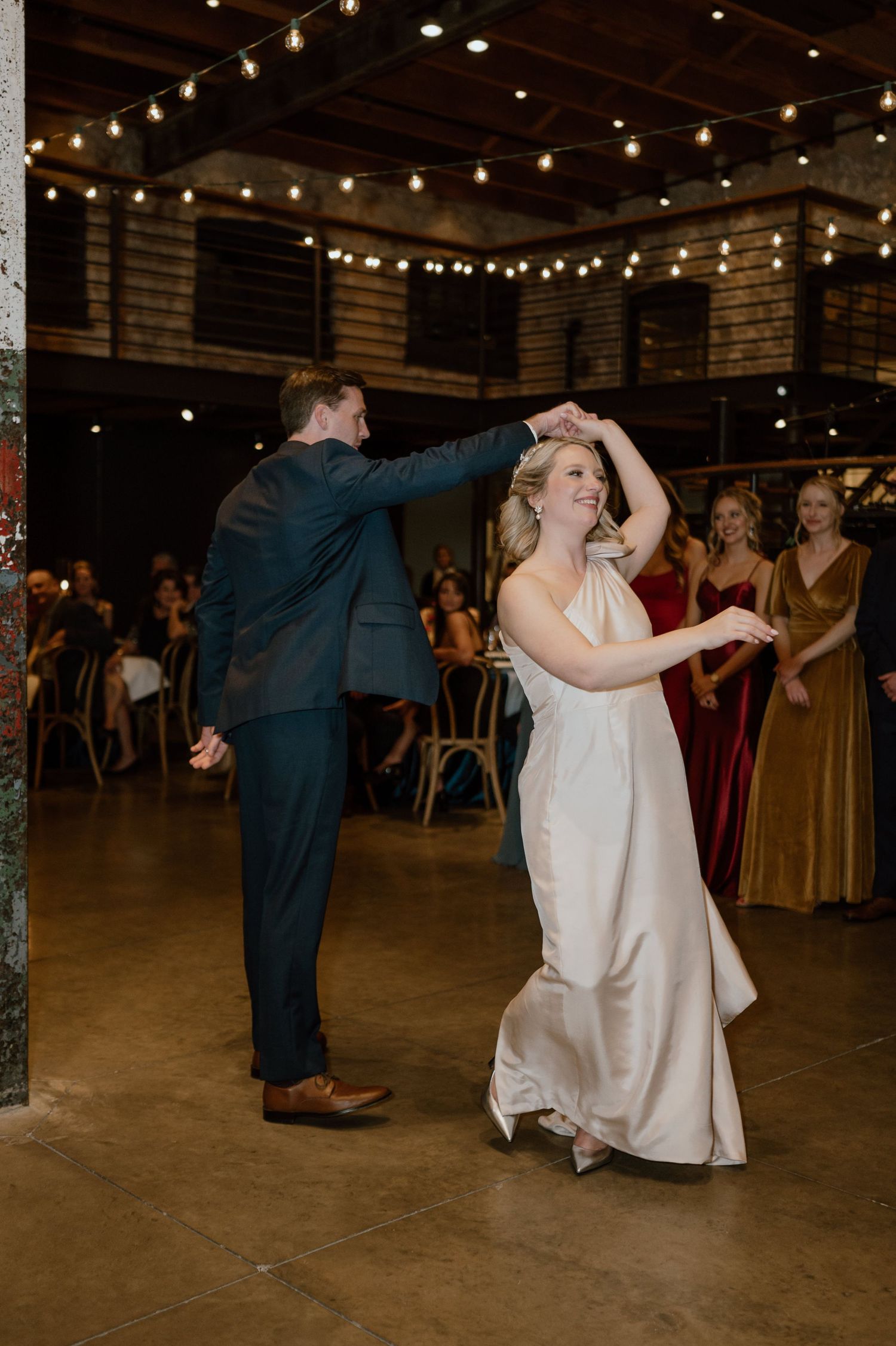 Wedding couple dancing and twirling in an industrial venue with string lights.