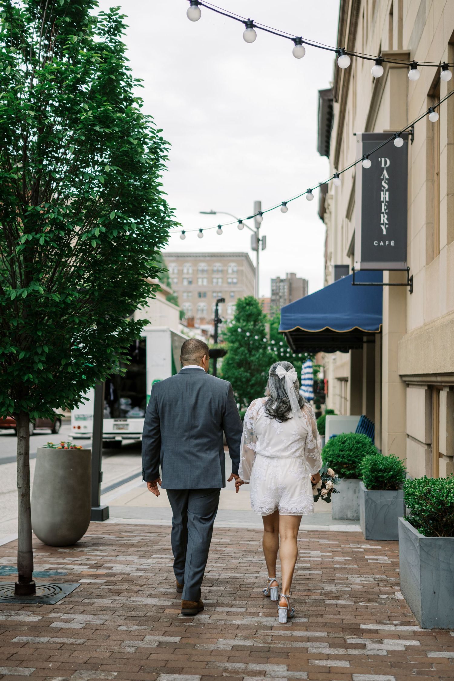 A couple walking down a charming city sidewalk with string lights and greenery overhead.