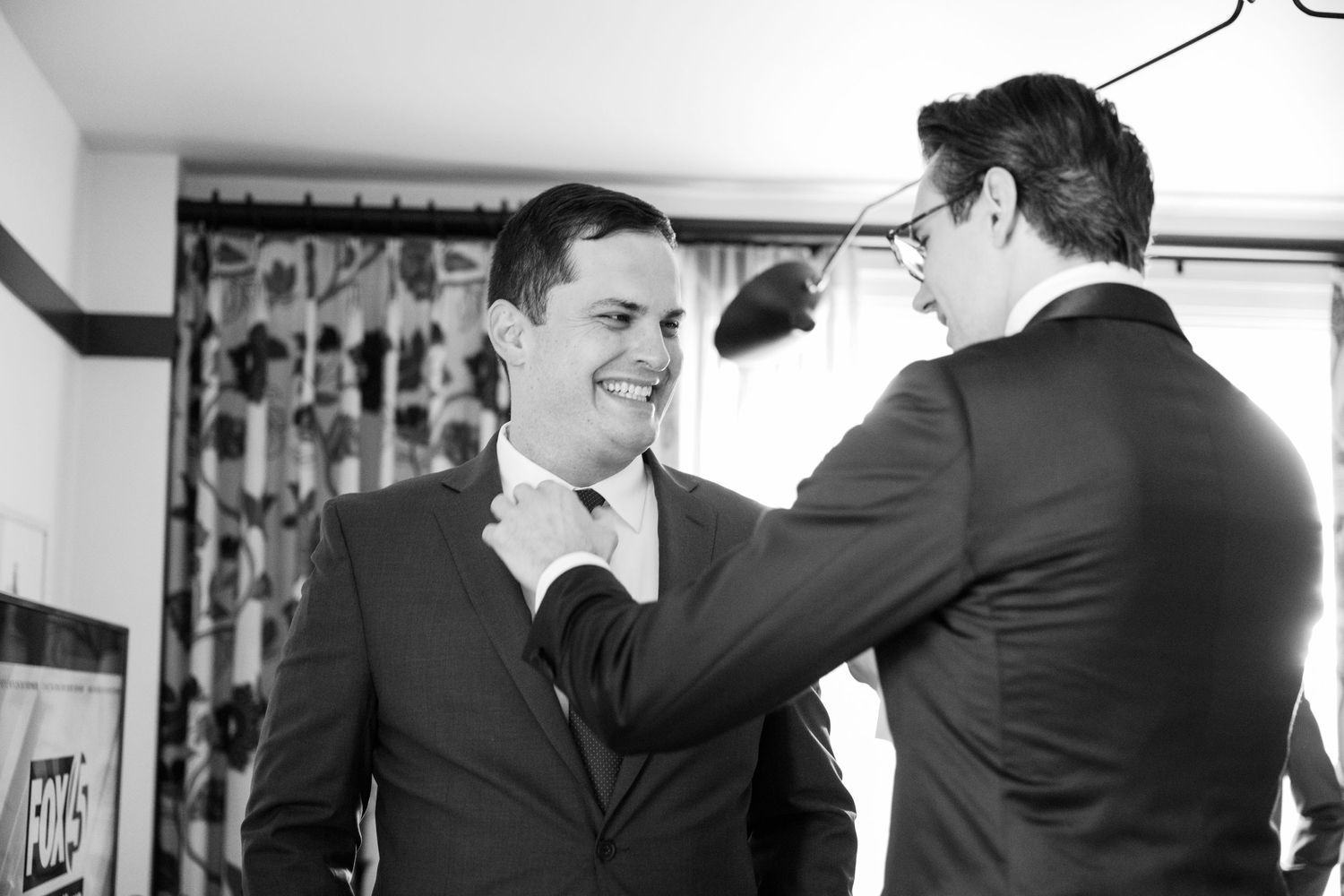 Black and white photo of wedding preparation moments in a room with floral curtains.