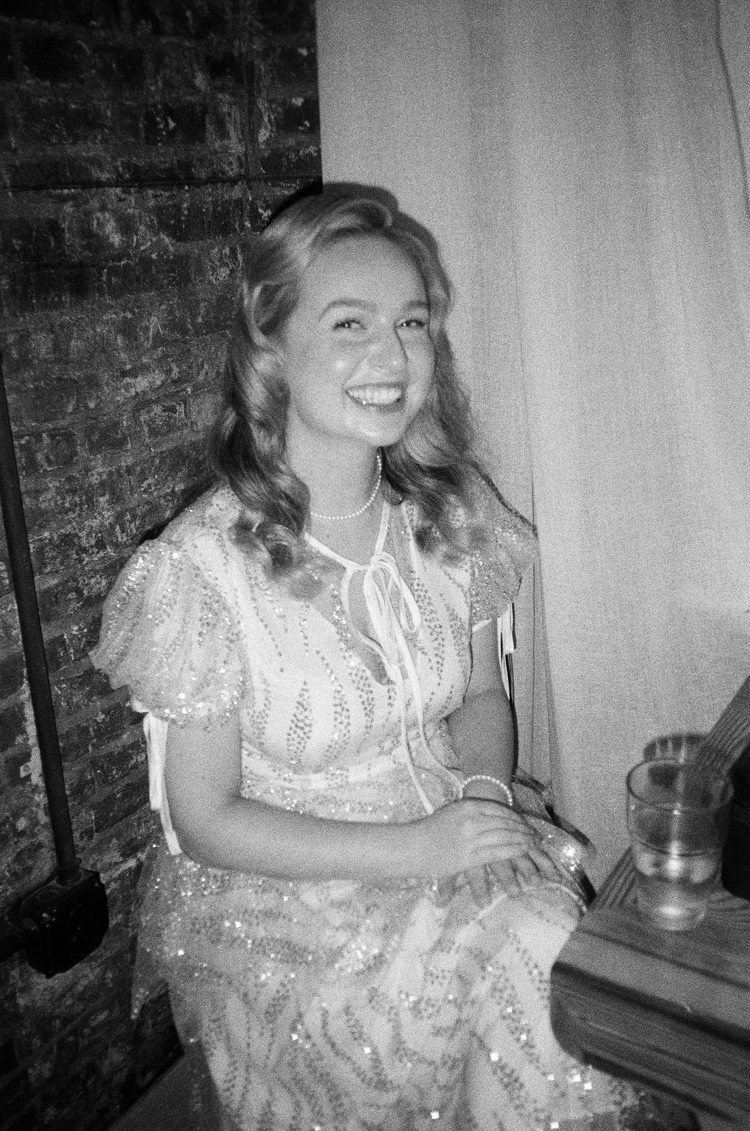 A woman in a vintage-style lace dress sits smiling in a rustic brick setting.