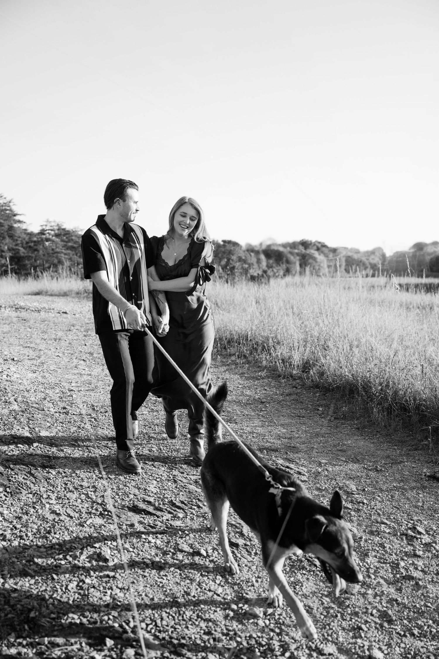 A couple walks their energetic dog on a dirt path through a scenic field on an overcast day.
