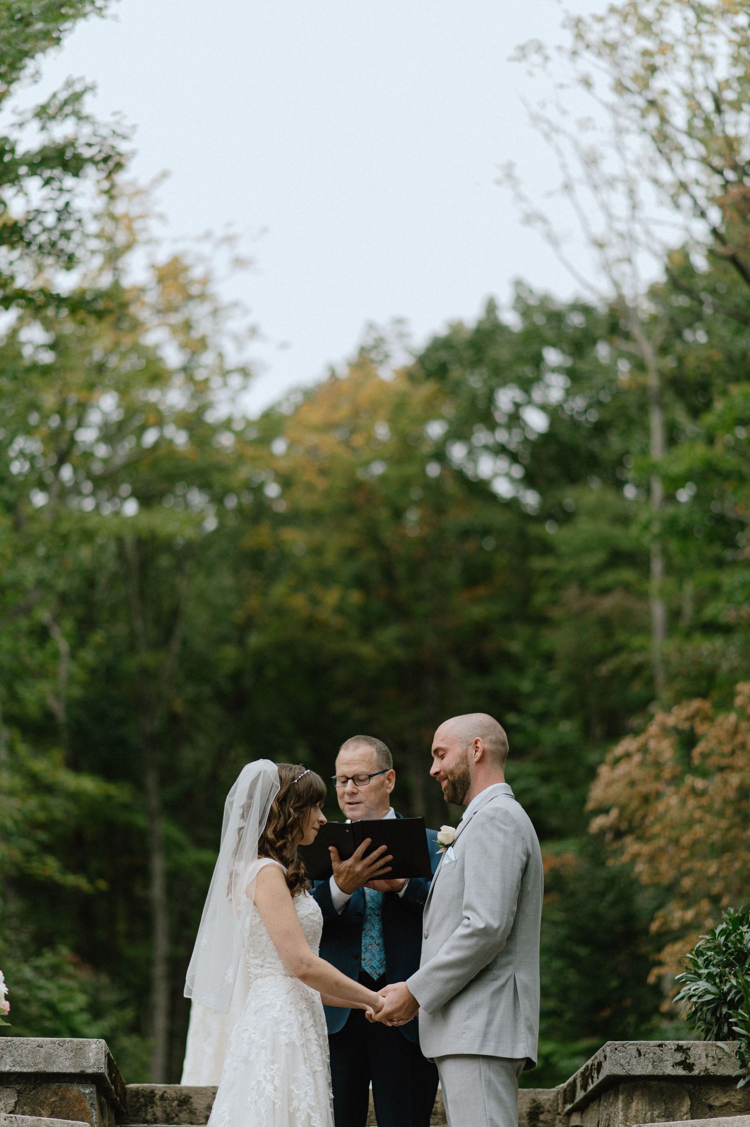 A wedding ceremony takes place on stone steps surrounded by lush greenery during an outdoor autumn wedding.