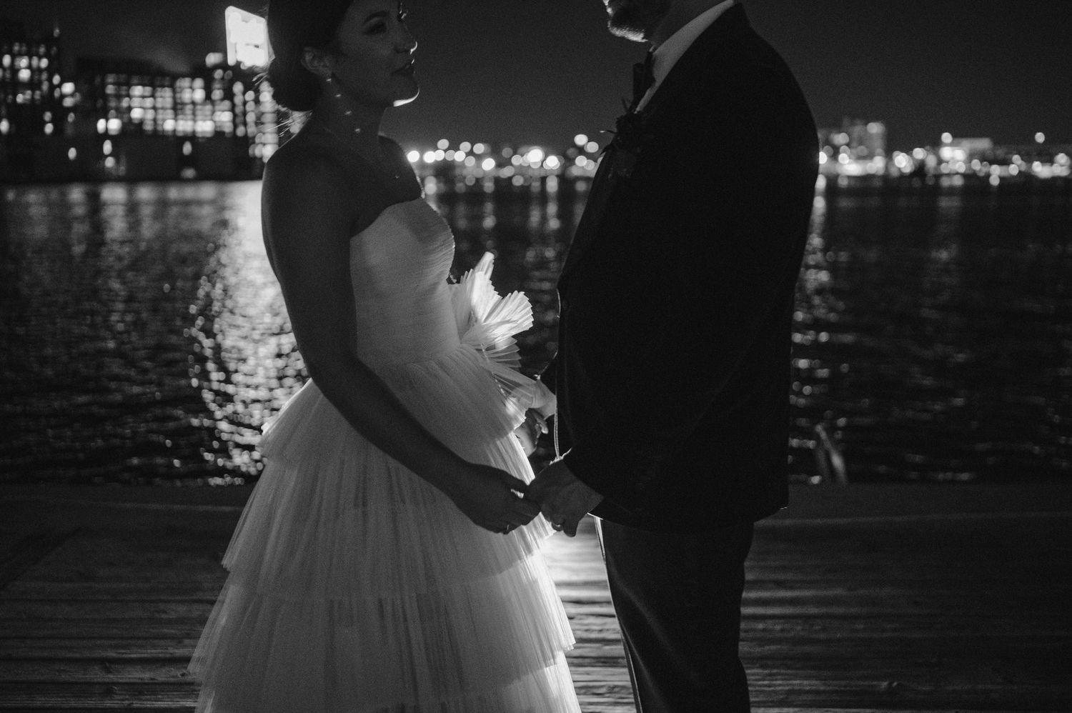 A romantic black and white portrait of newlyweds by the waterfront with city lights in the background.