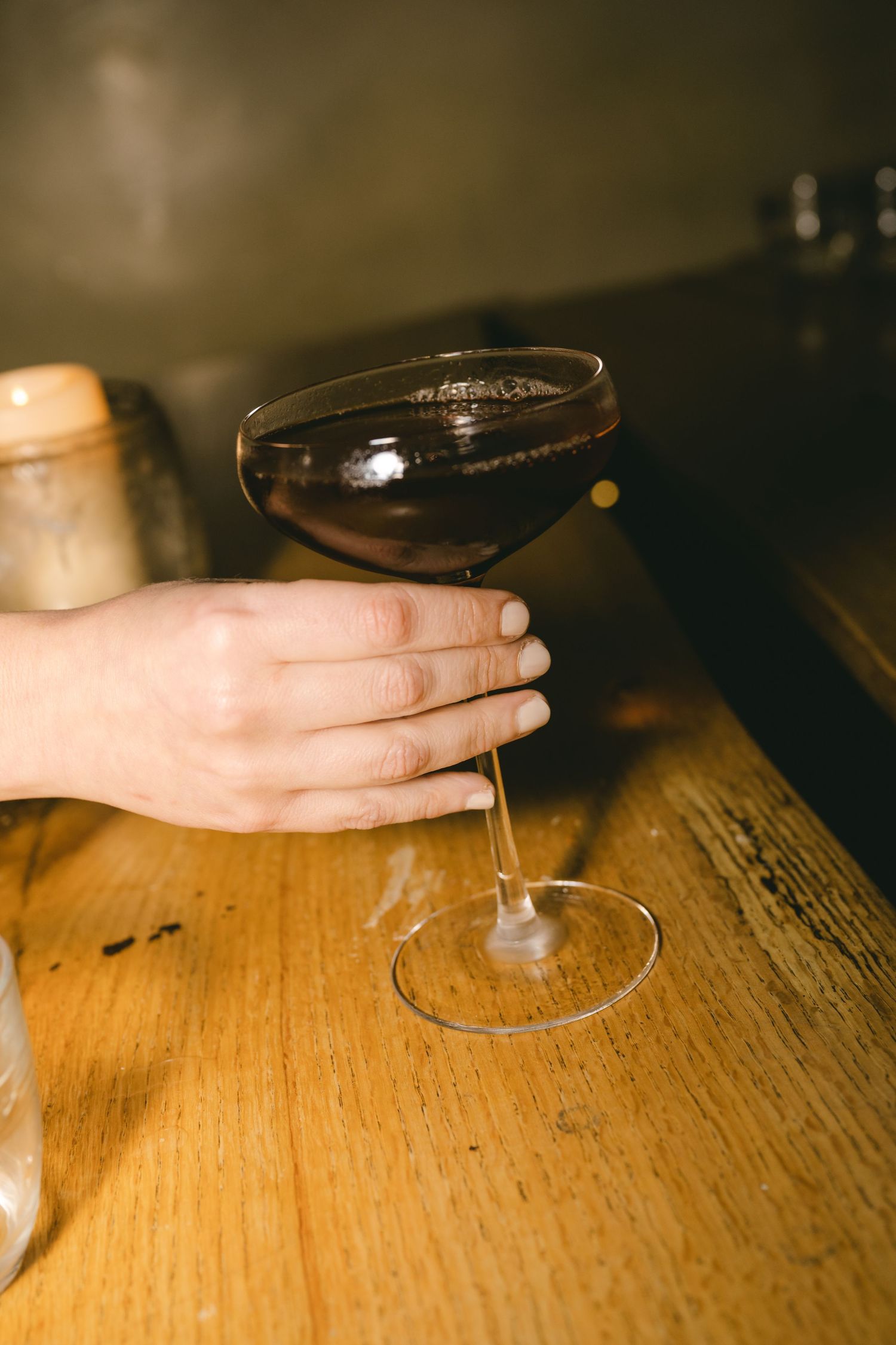 A hand holds a dark cocktail in a coupe glass on a wooden surface in moody lighting.