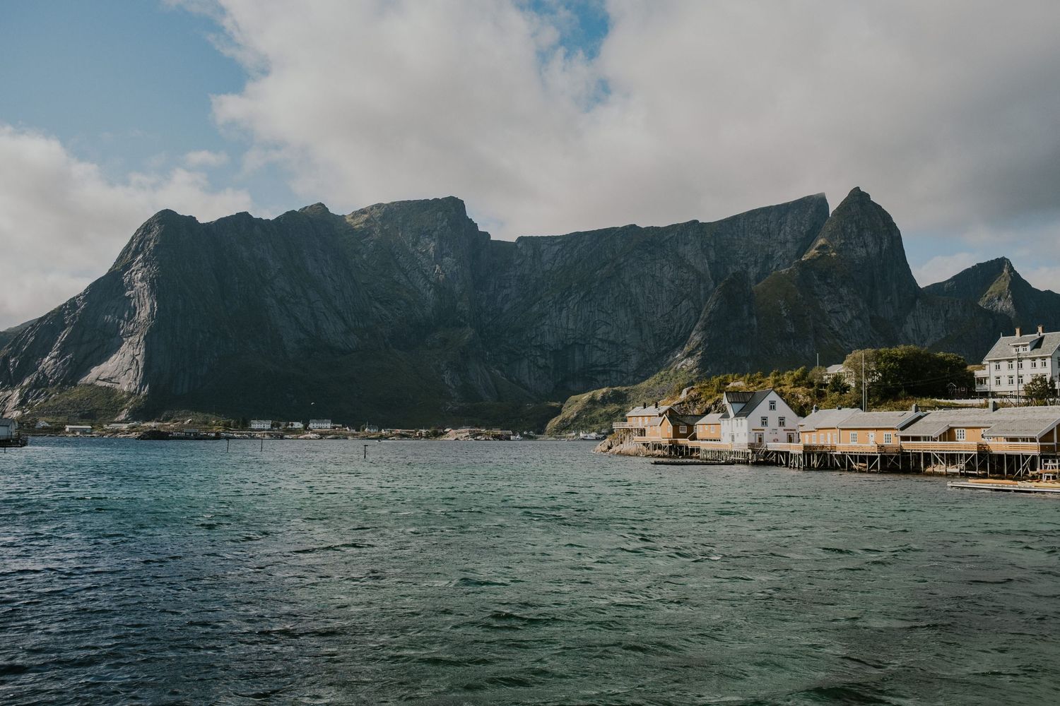 Rugged coastal mountains tower over small fishing village with waterfront buildings in Norwegian fjord.