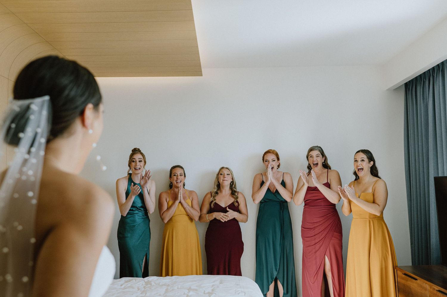 Bridesmaids in colorful dresses react to seeing the bride for the first time.
