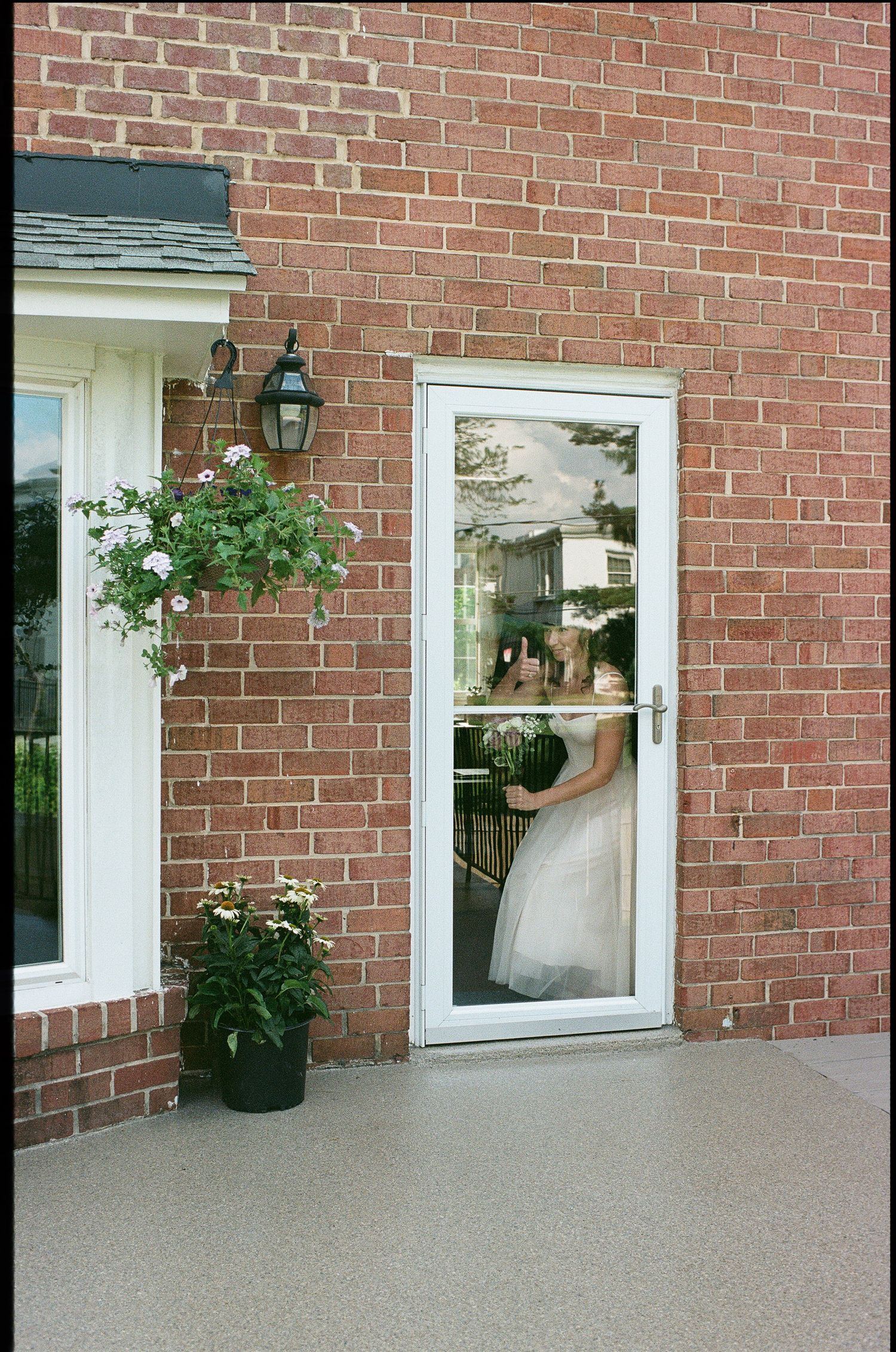 A white storm door with a reflection of a bridal dress next to potted flowers against a brick house exterior.