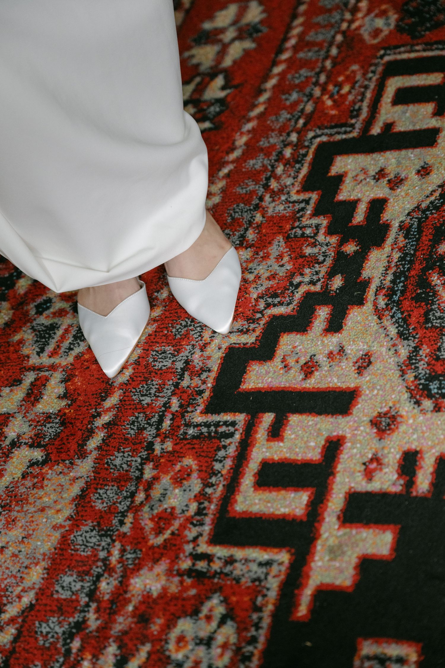 White shoes standing on an ornate red and black patterned carpet.