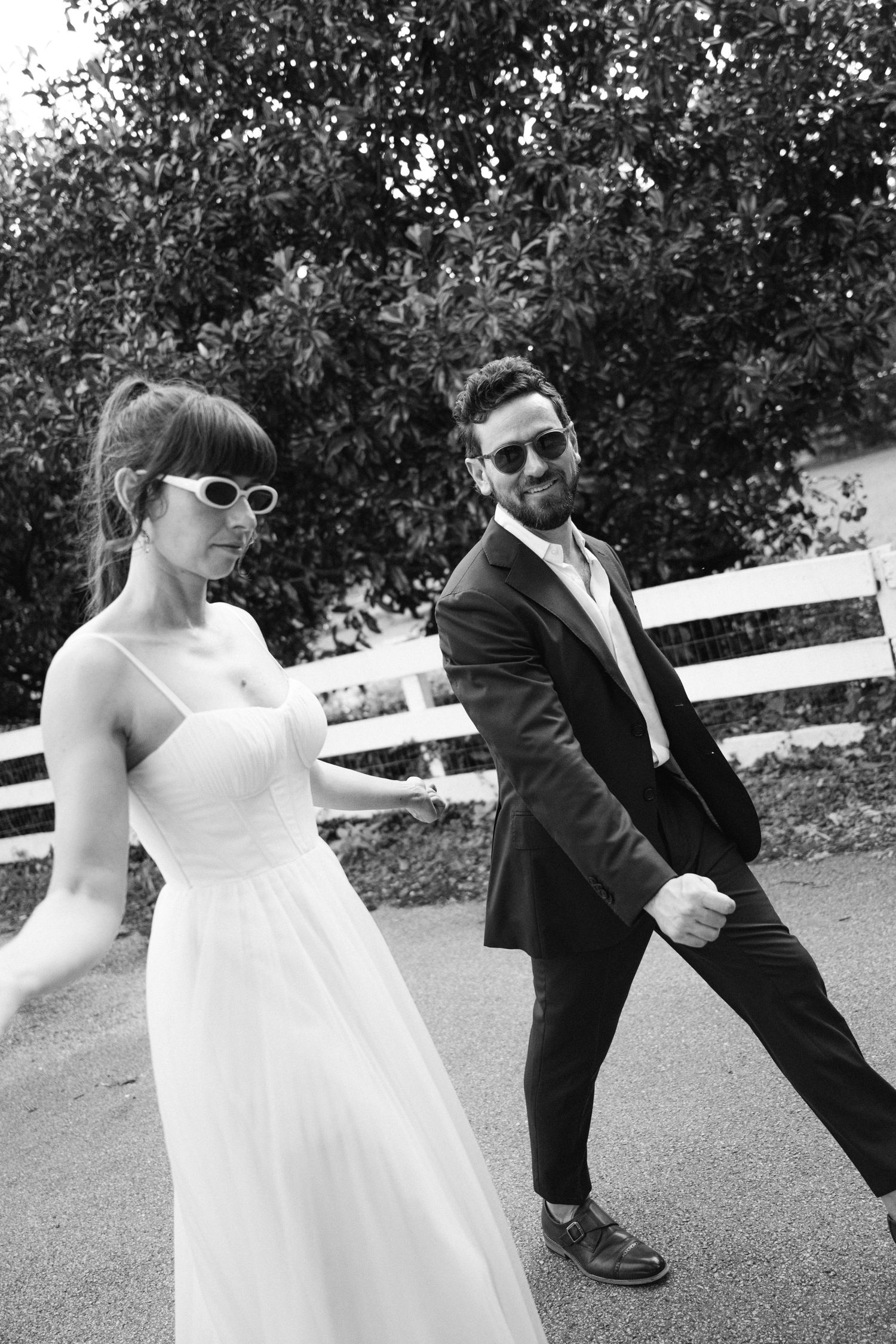 Playful wedding couple poses dramatically against white fence in black and white photography.