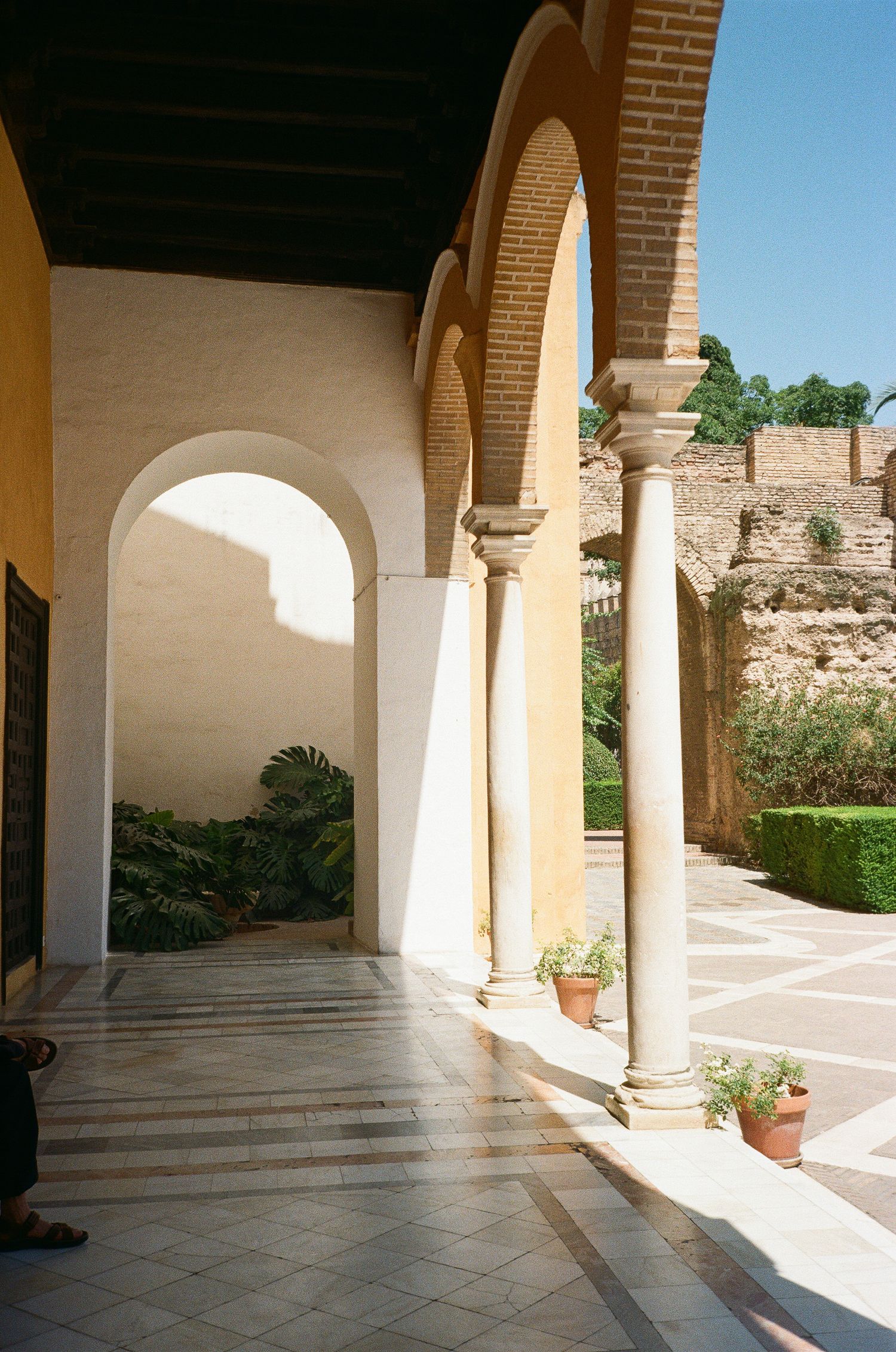 Sunlit colonnade with white arched columns and brick walls creating dramatic shadows on tiled floor.
