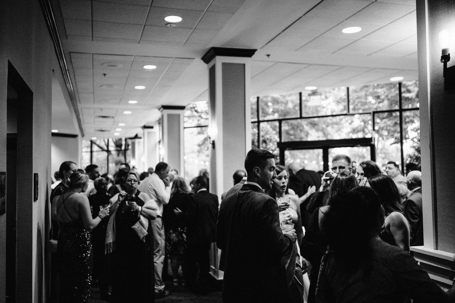 A black and white photo of wedding guests mingling in a reception hall with columns.