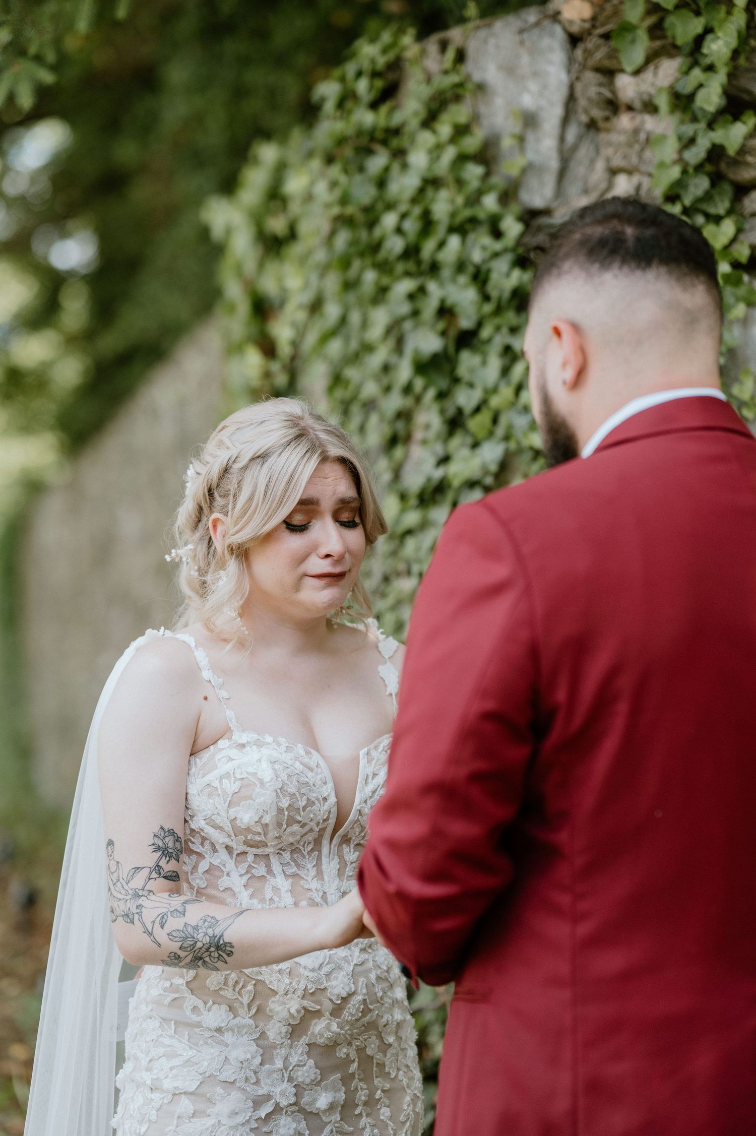 Wedding couple standing together against an ivy-covered wall with bride in lace dress and groom in red suit.