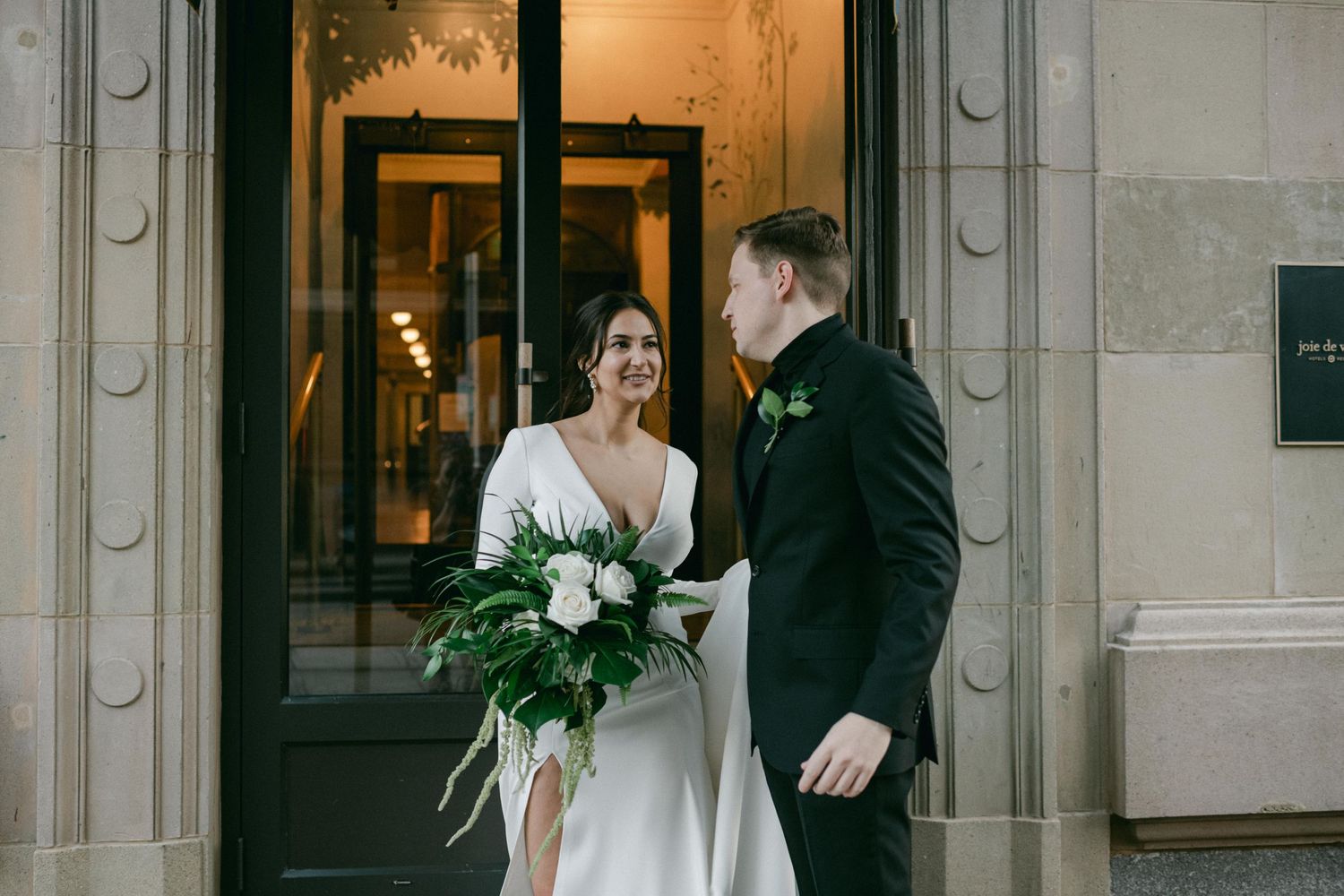 Elegant wedding couple sharing a moment outside a grand building entrance with lush green bouquet.