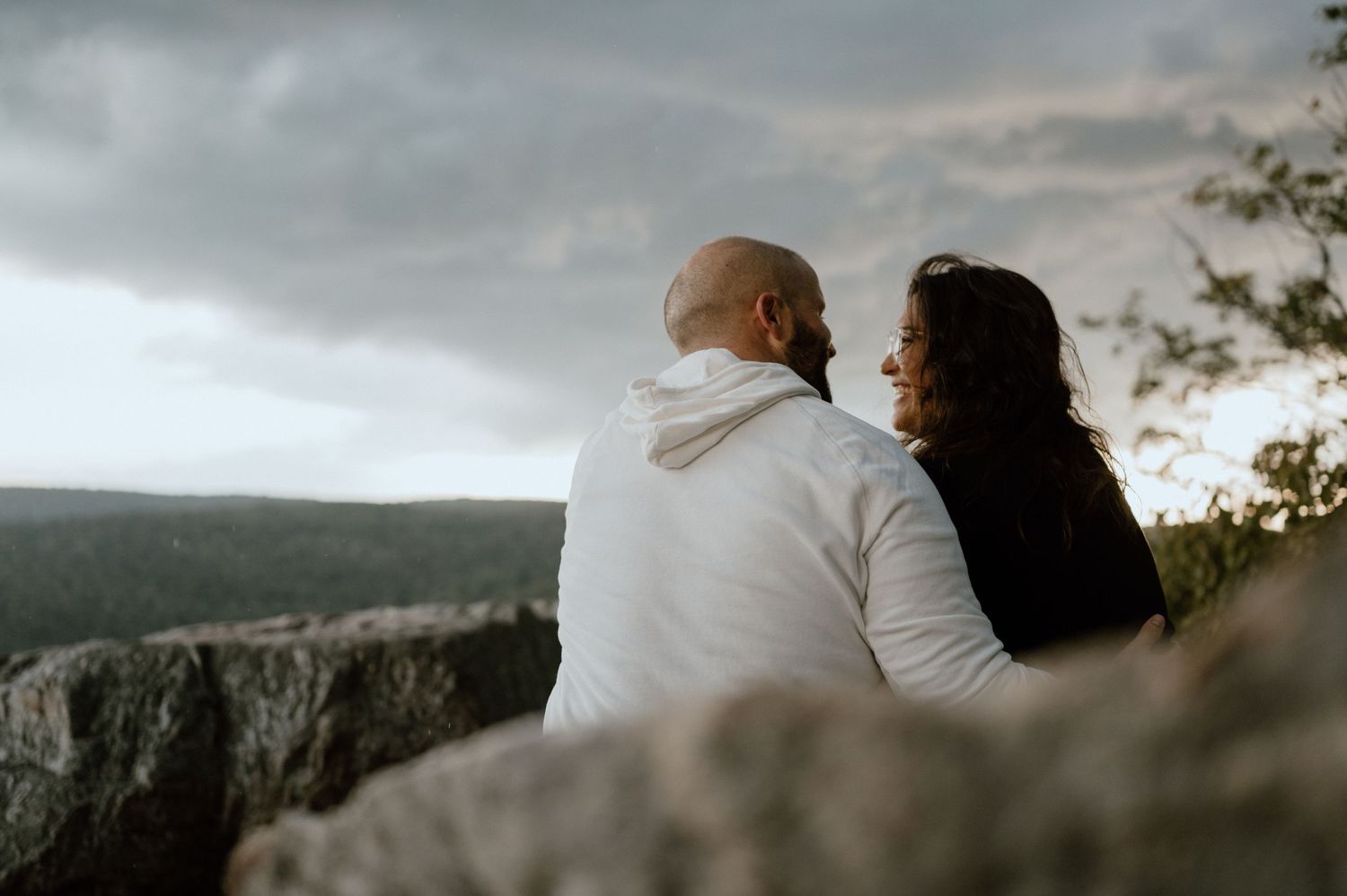 A couple sitting close together on rocky cliff edge overlooking misty mountain vista.