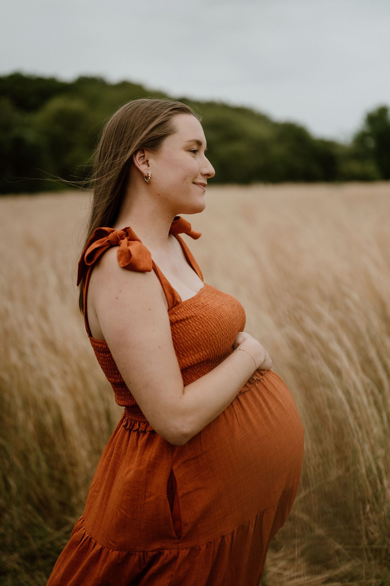 A maternity photo featuring a rust-colored dress against tall grasses and cloudy skies.