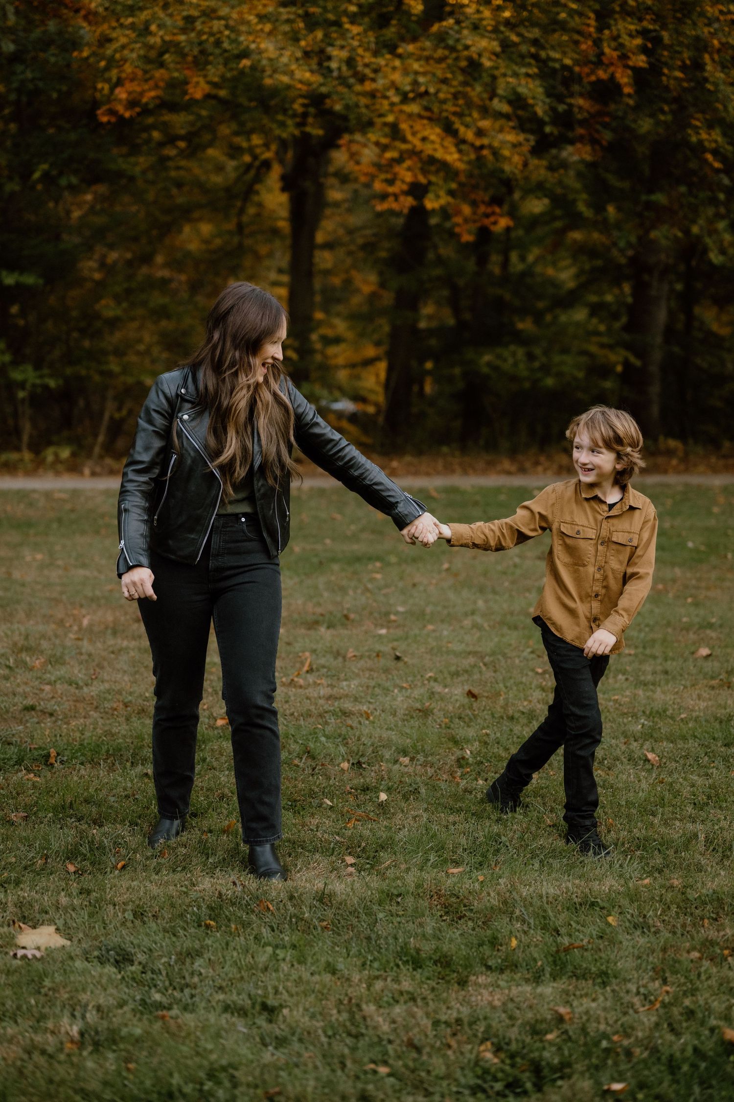 Two people hold hands while standing in a grassy field surrounded by autumn trees.