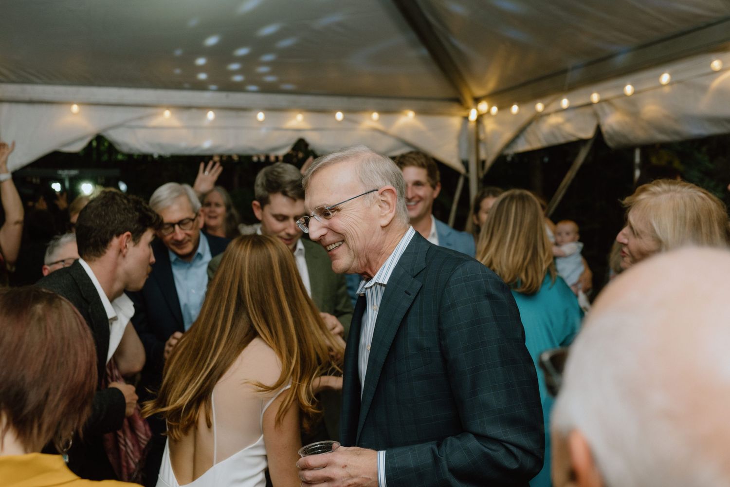 Guests mingle and socialize during an evening wedding reception under a tent with twinkle lights.
