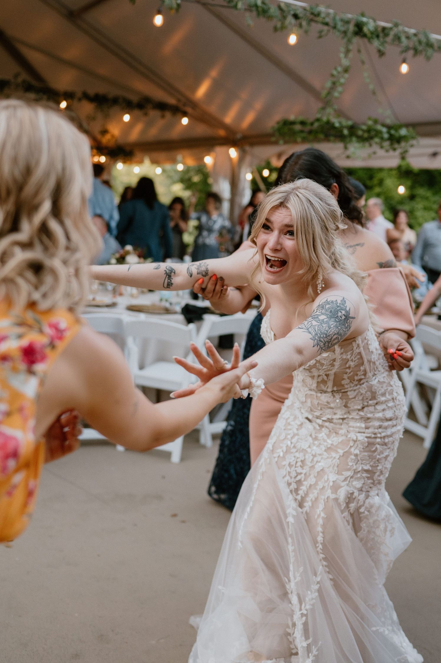 Joyful bride in a lace dress dances with a wedding guest under a tent with string lights and greenery.