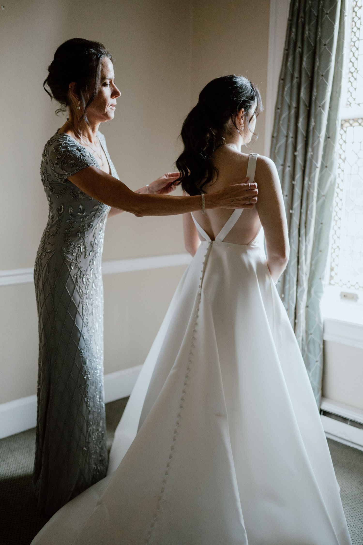 A tender moment as someone in a silver dress helps adjust a classic white wedding gown by a window.