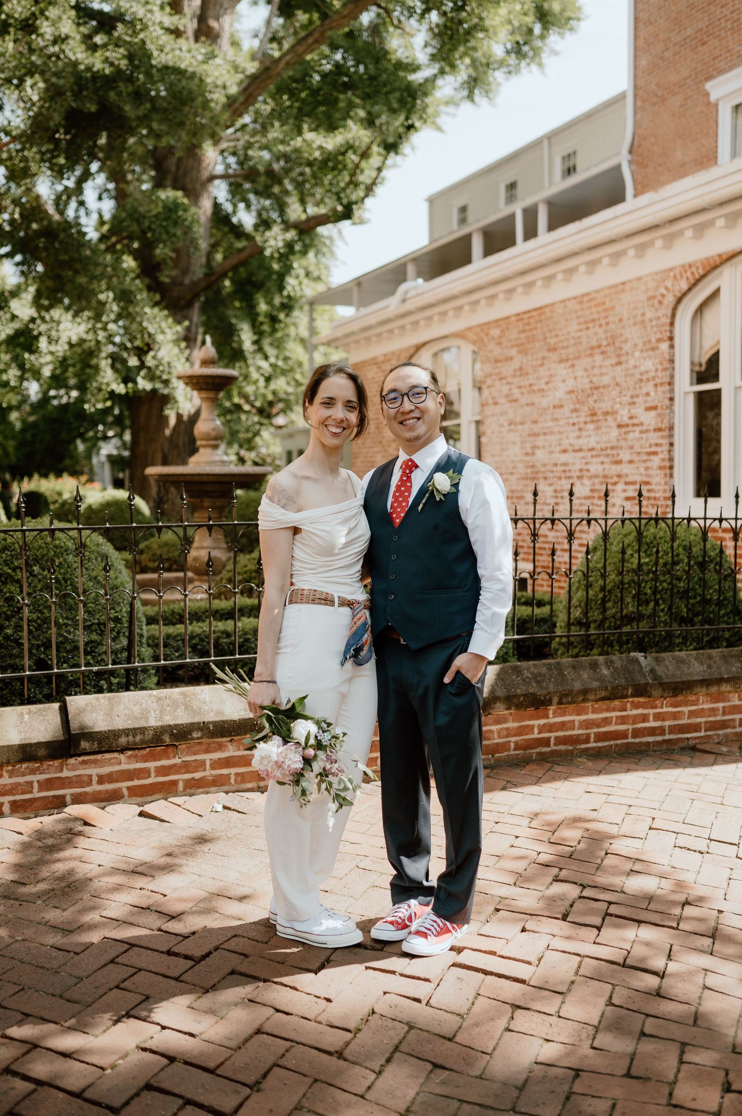 A couple in wedding attire poses on a brick patio with trees and architecture in the background.
