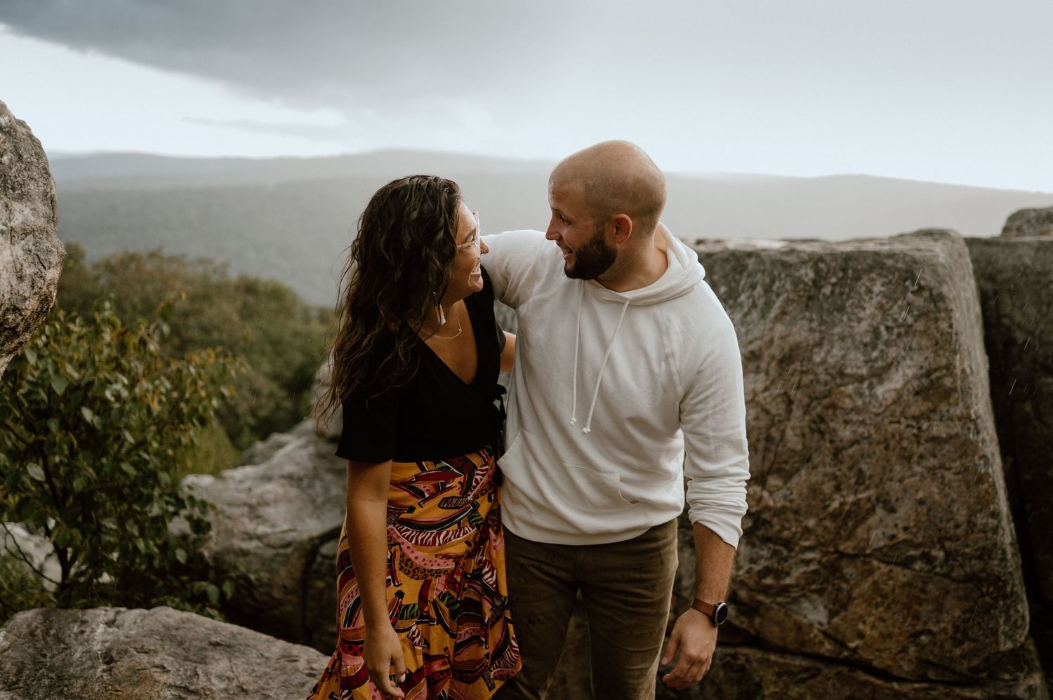 Couple in casual attire standing close together on rocky mountain ledge.