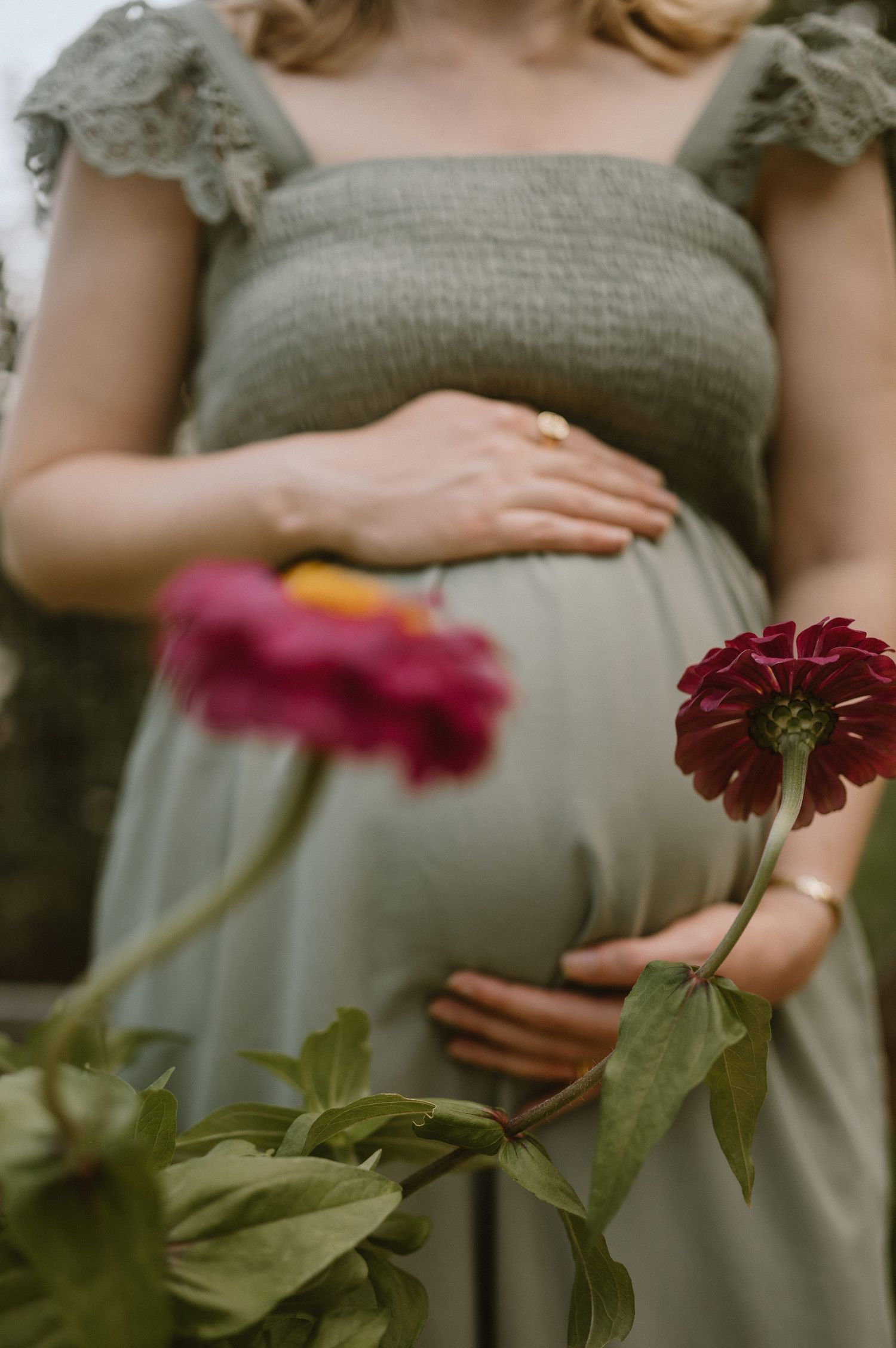 A close-up of hands resting on a sage green dress with pink zinnias in the foreground.
