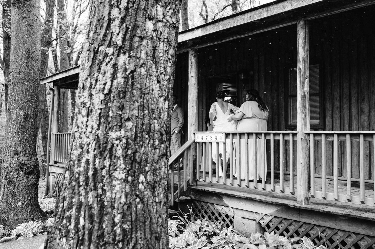 A vintage black and white photograph shows a rustic cabin porch with figures standing behind the railing.