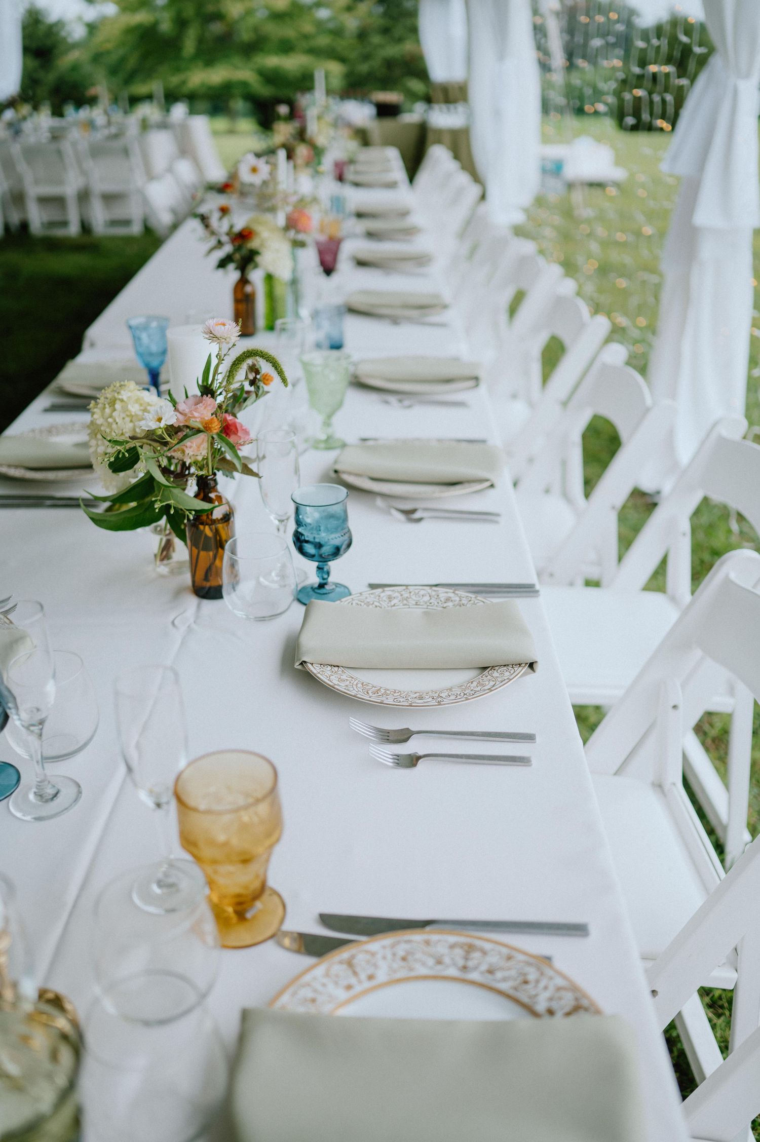 An elegant outdoor wedding reception table with white chairs, floral centerpieces and blue glassware.
