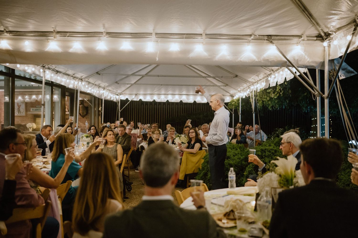 Speaker addressing crowd during evening reception under white tent with string lights.