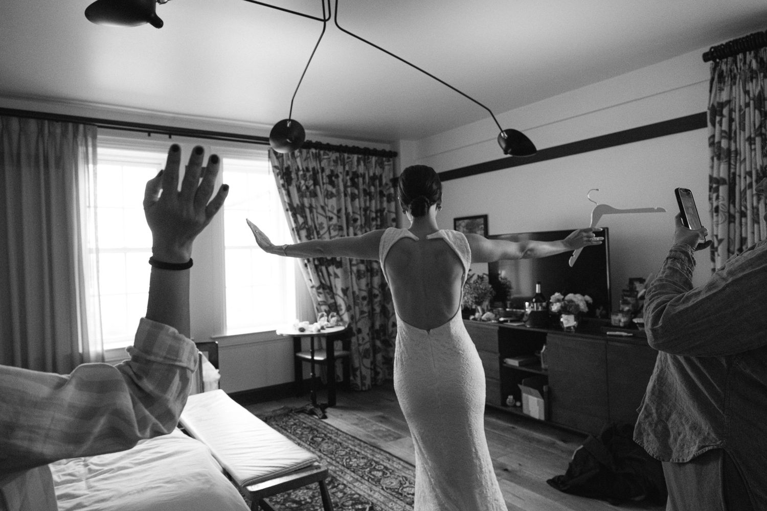 A black and white candid moment captures wedding preparations in a sunlit room with floral curtains.