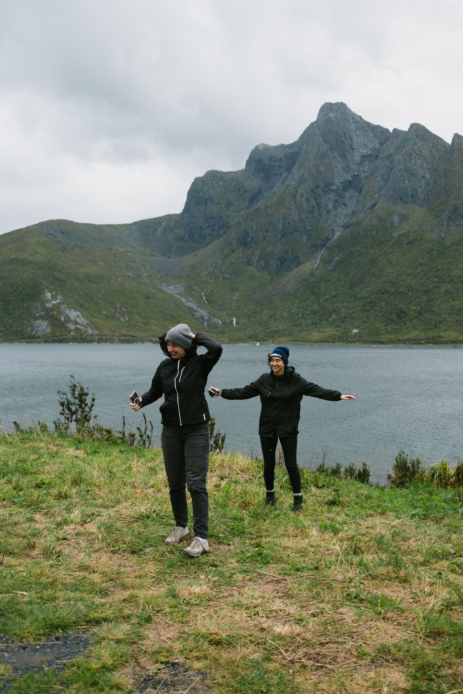 Two people dancing or jumping joyfully by mountain lake with dramatic peaks in background.