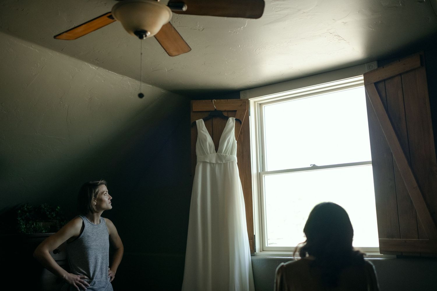 White wedding gown hanging by window as people gather in preparation room.