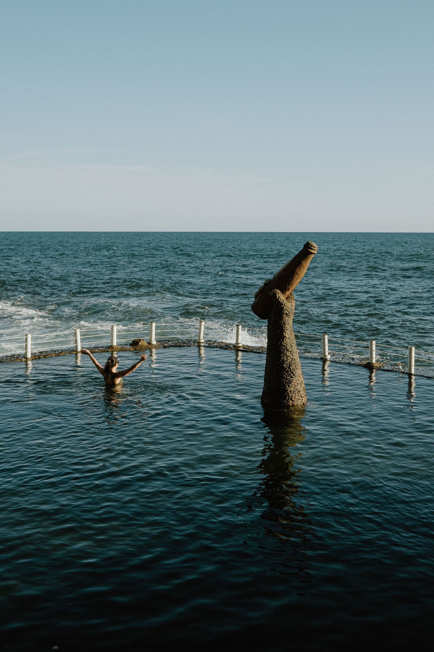 Ocean pool with sculptural post protruding from water and swimmers enjoying the calm waters against ocean backdrop.