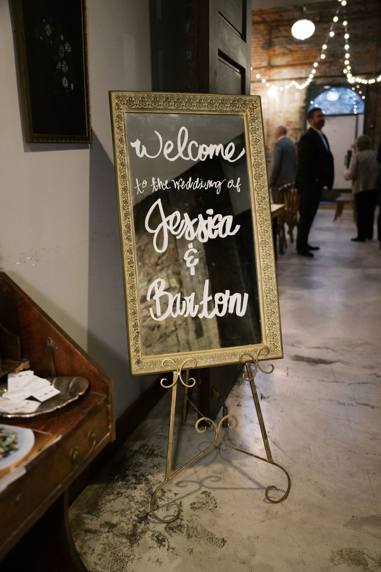 Gold-framed welcome sign on an easel displays handwritten text for a wedding reception in a rustic indoor venue.