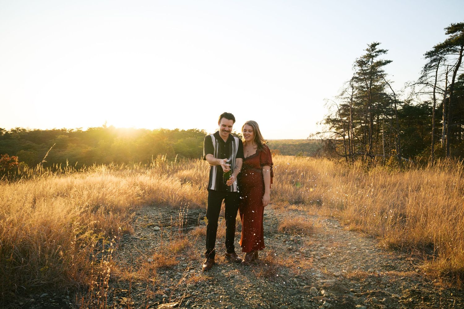 A couple stands together in a golden field during sunset with evergreen trees in the background.