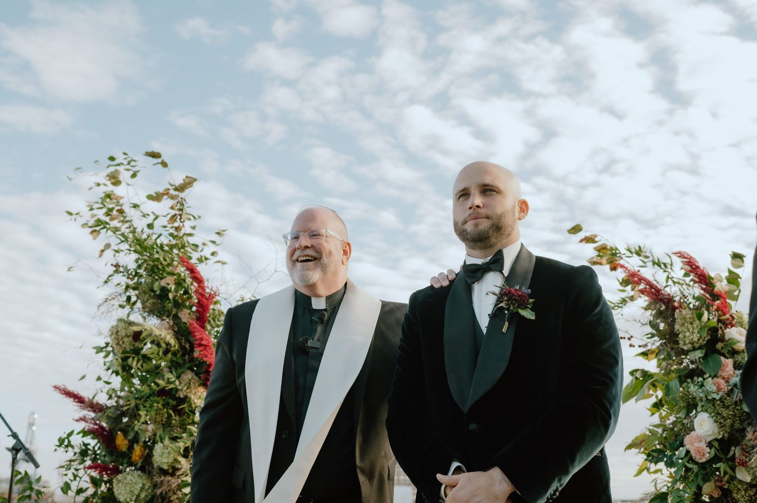 Two people in formal attire stand at an outdoor wedding altar decorated with red floral arrangements.