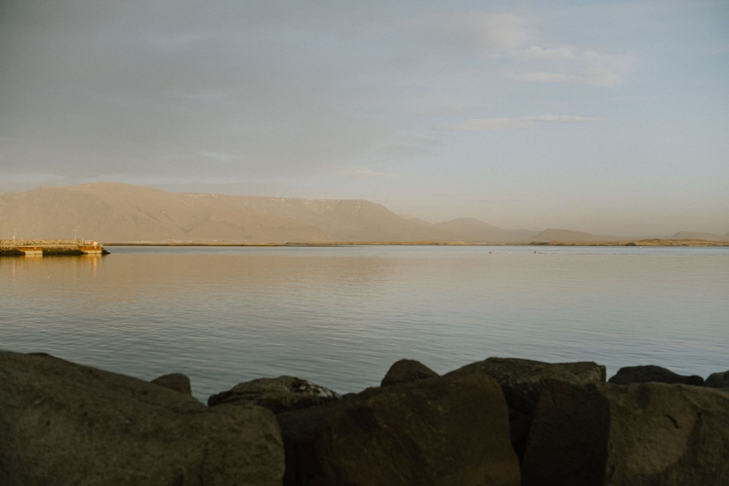 Rocky shoreline at sunset with mountains visible across calm waters.