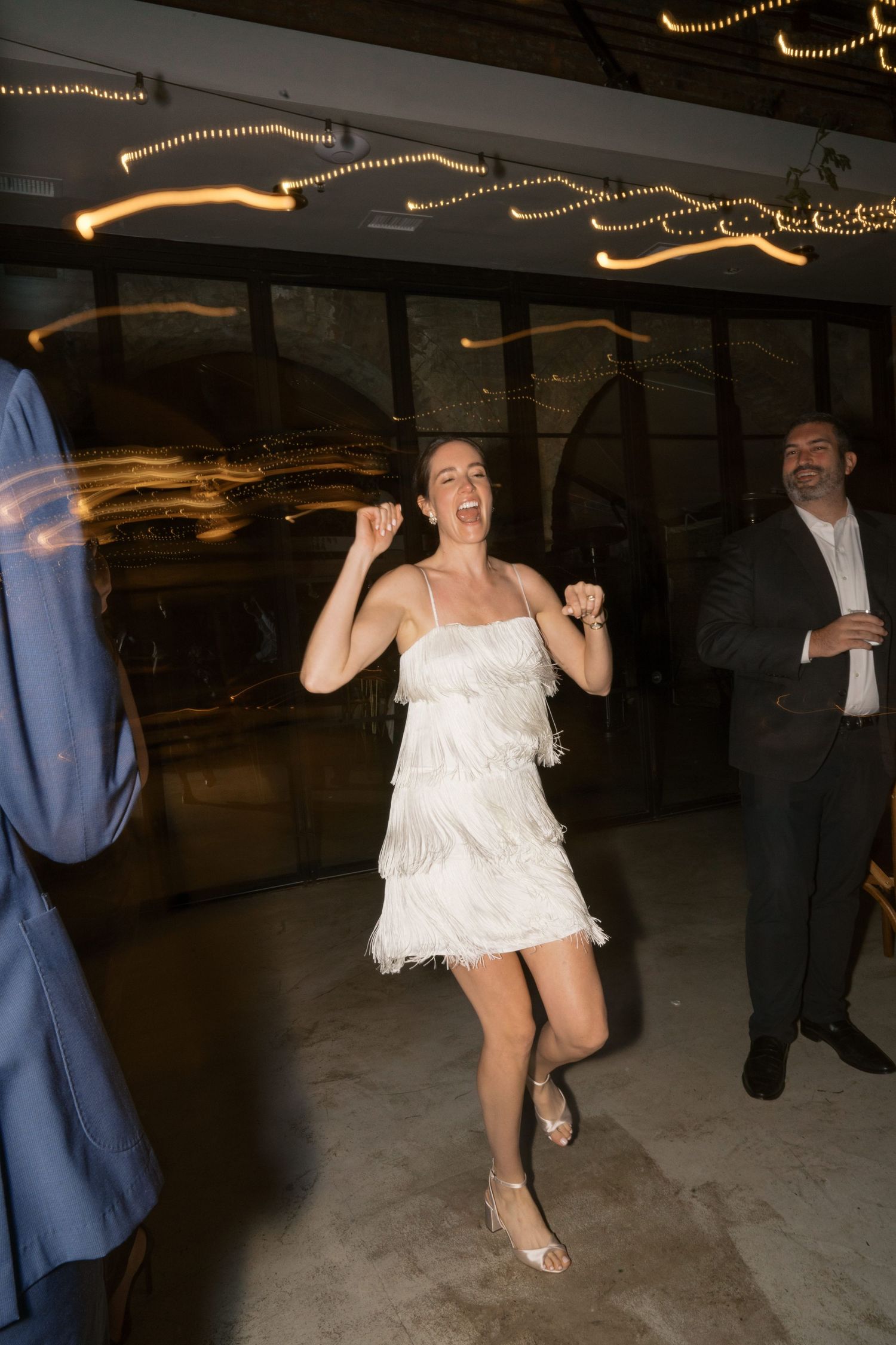 A celebratory dance scene with a woman in a white fringe dress dancing energetically under string lights.