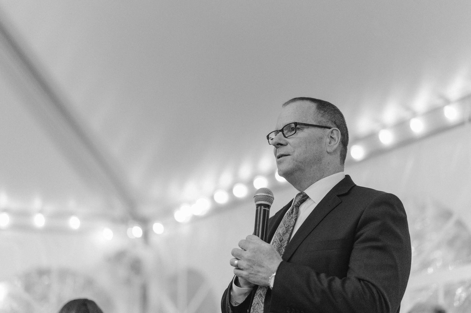 Speaker holding microphone during wedding reception under white tent.