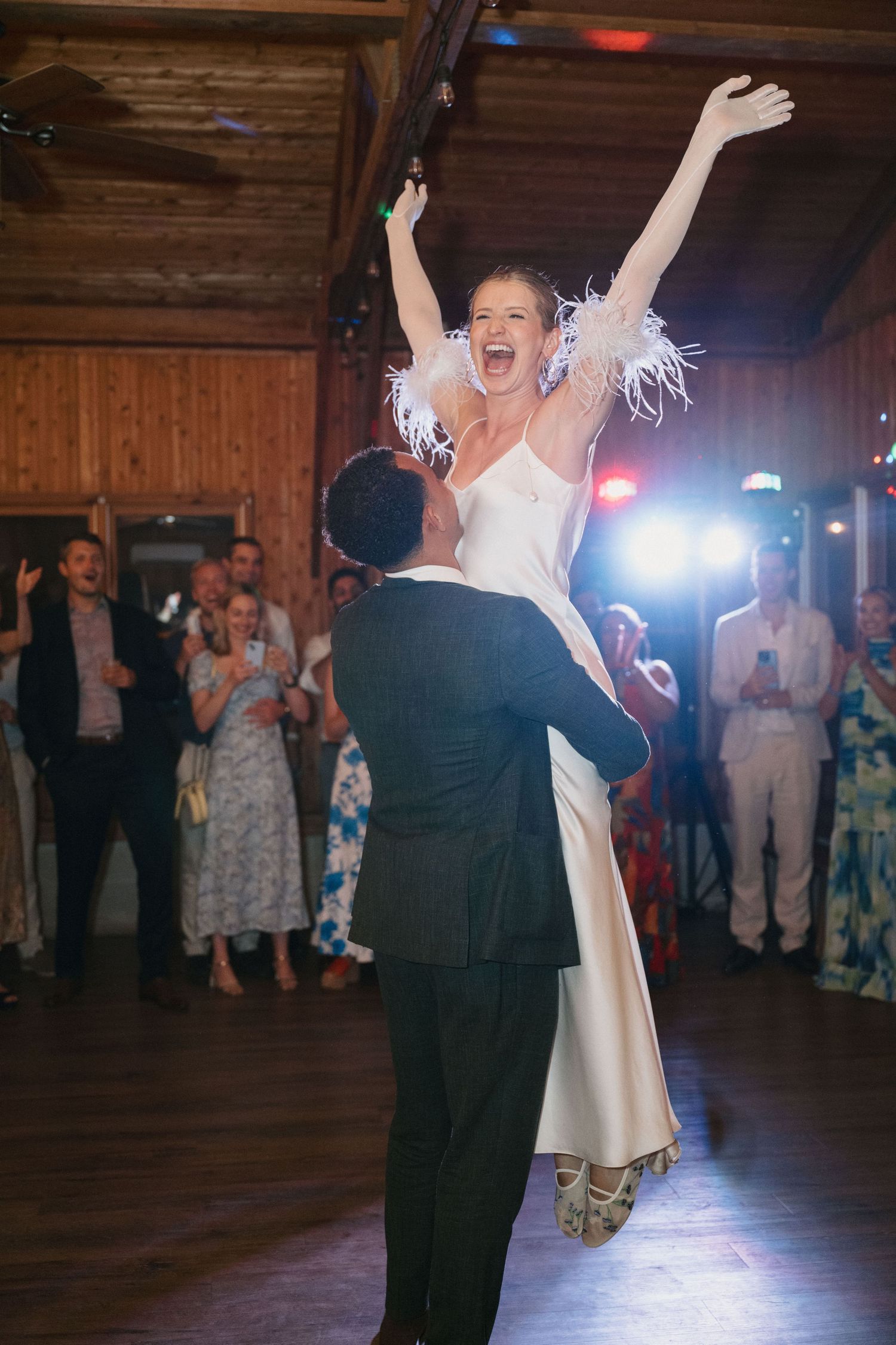 A bride in a white dress is lifted joyfully during a celebratory dance at a wedding reception.