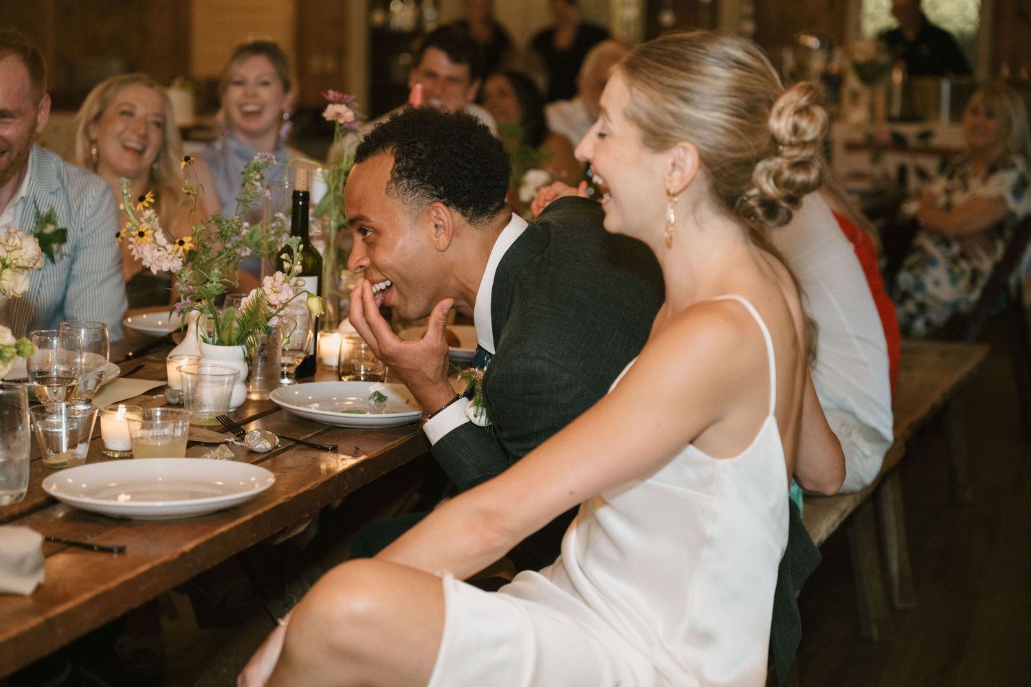 Wedding reception guests laughing and enjoying dinner at a rustic wooden table.