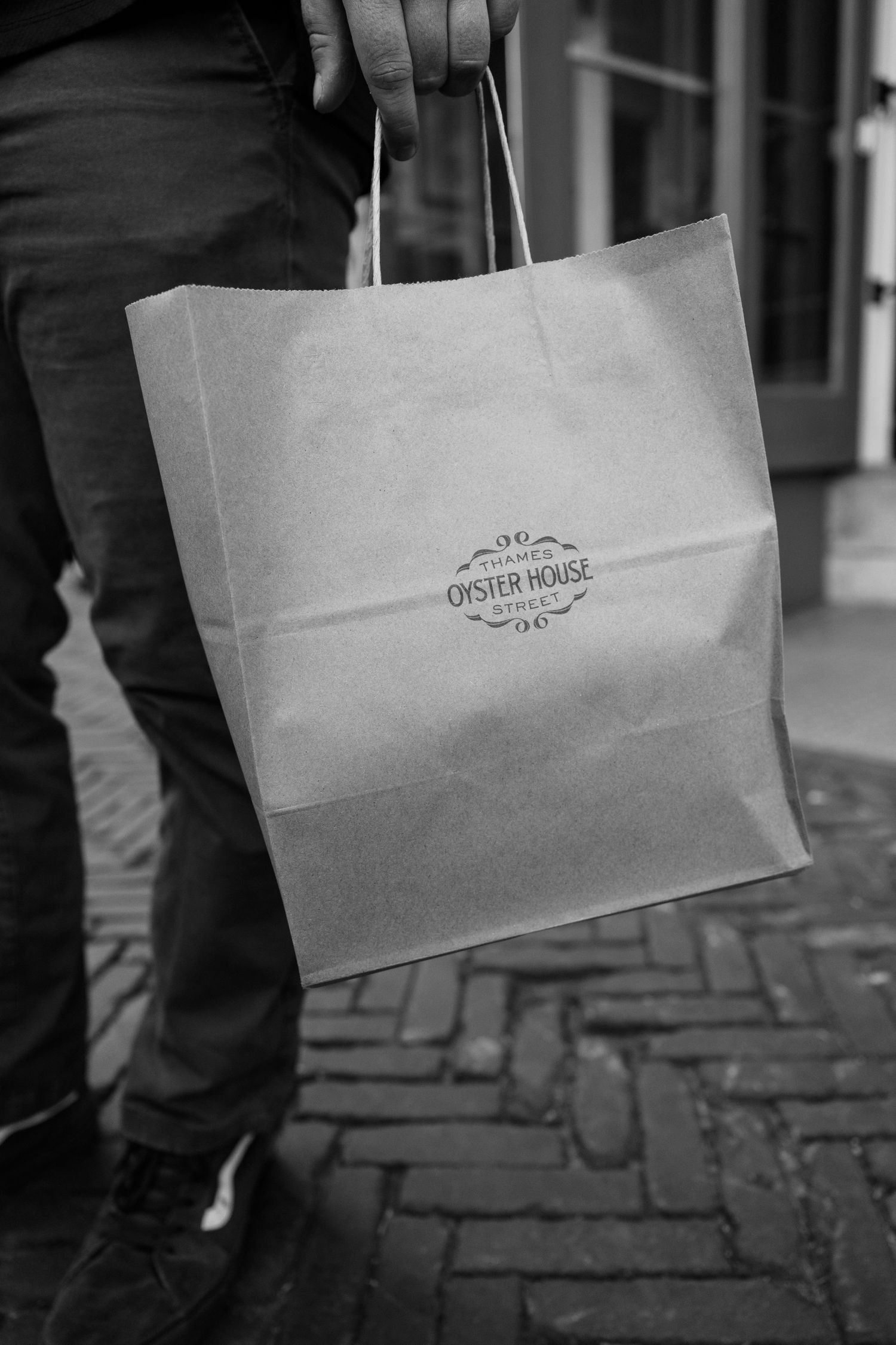 A person holding a white shopping bag with an ornate logo stands on a brick sidewalk in black and white.