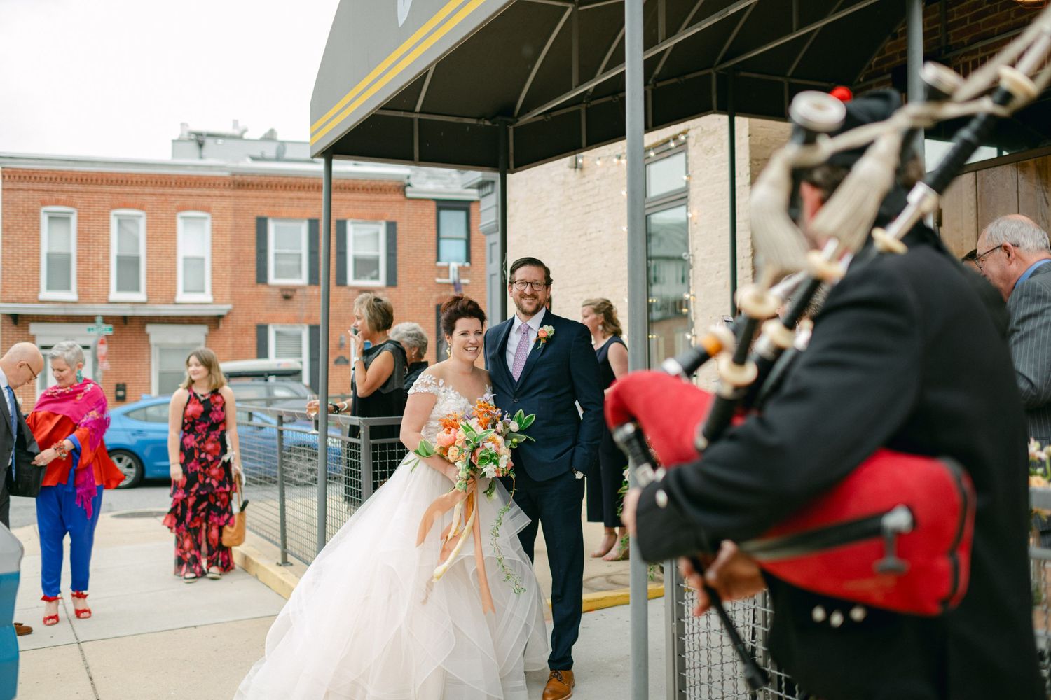A wedding party gathers outside a brick building entrance with guests looking on and photographers capturing the moment.
