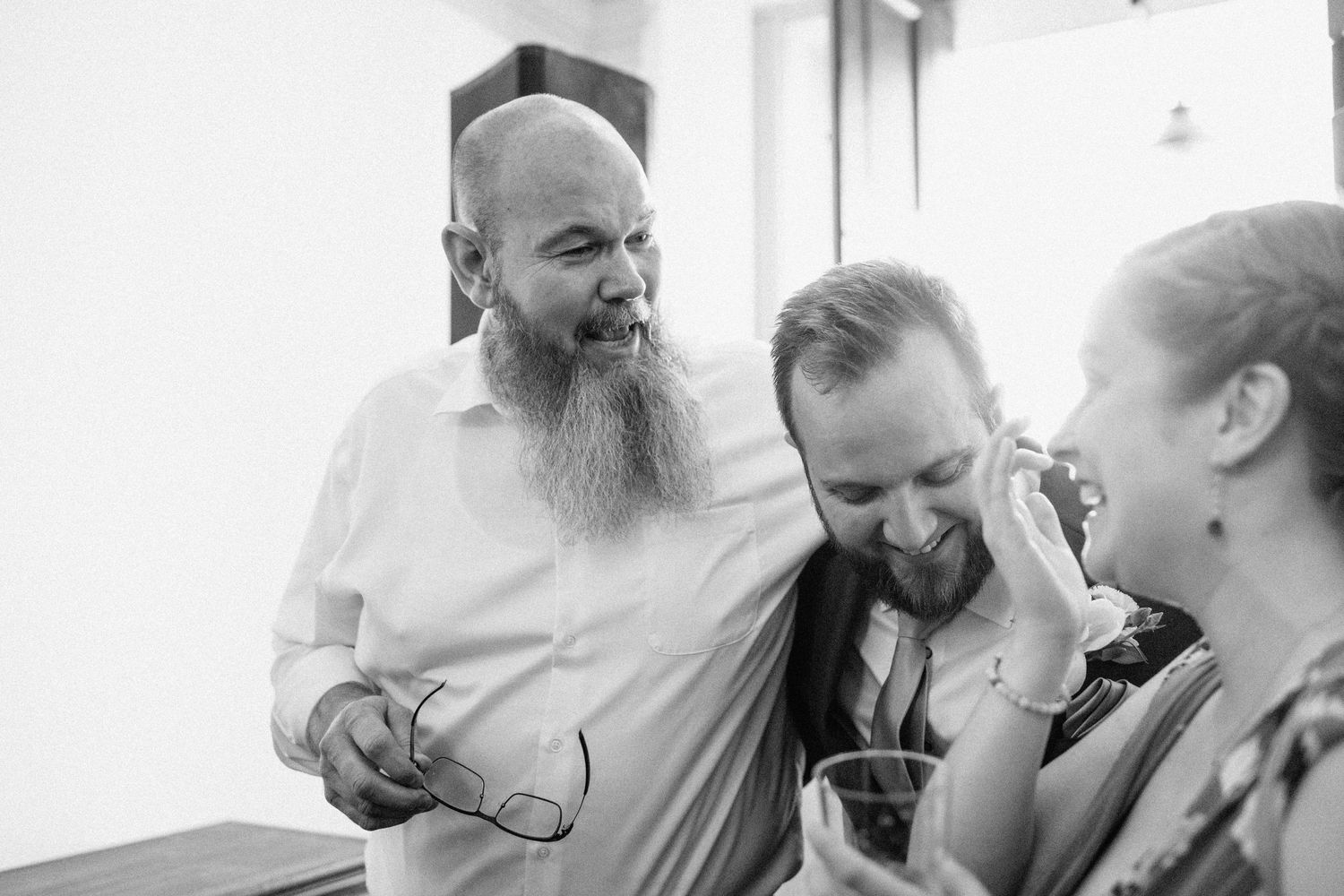 Black and white candid photo showing a bearded man and others laughing together during a wedding celebration.